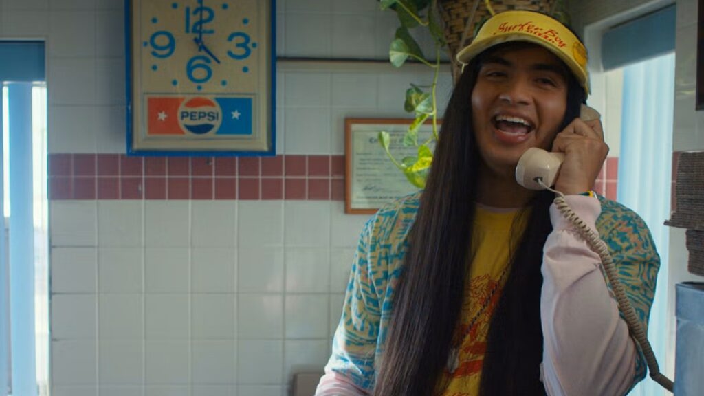 Argyle (Eduardo Franco) wearing a yellow cap and colorful shirt, talking on a corded phone in a kitchen with a bright, cheerful expression and 1980s decor including a Pepsi clock.