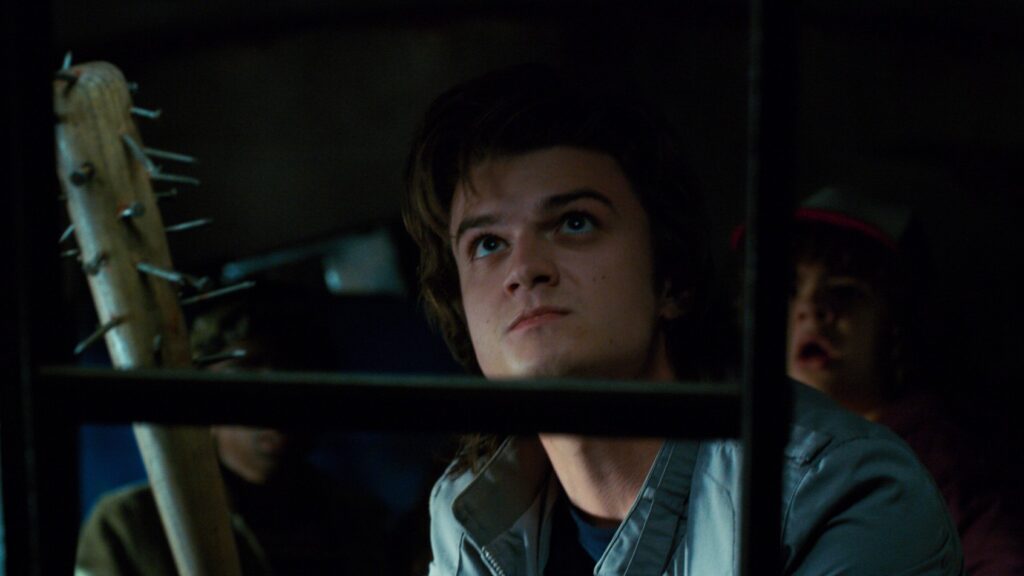 Steve Harrington (Joe Keery) looking upward with a concerned expression, photographed from below in dim lighting, holding a spiked club, and a blurred Dustin Henderson (Gaten Matarazzo) is visible in the background.