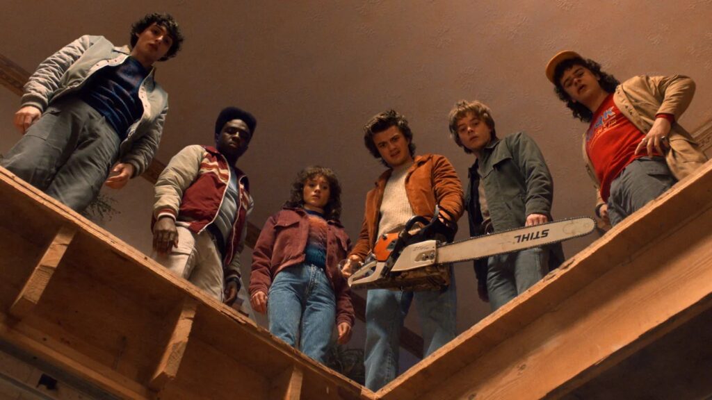 Mike Wheeler (Finn Wolfhard), Lucas Sinclair (Caleb McLaughlin), Nancy Wheeler (Natalia Dyer), Steve Harrington (Joe Keery), Jonathan Byers (Charlie Heaton), and Dustin Henderson (Gaten Matarazzo) standing on wooden stairs looking down, with Steve Harrington holding a chainsaw.