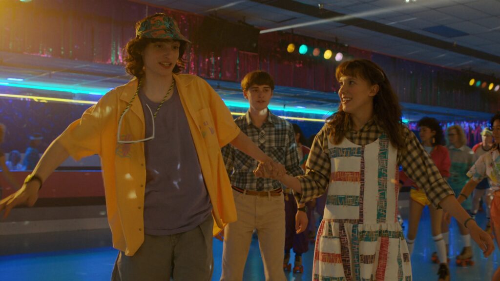 Mike Wheeler (Finn Wolfhard) in a bucket hat dancing with Eleven (Millie Bobby Brown), both smiling on a skating rink, while Will Byers (Noah Schnapp) is visible in the background.
