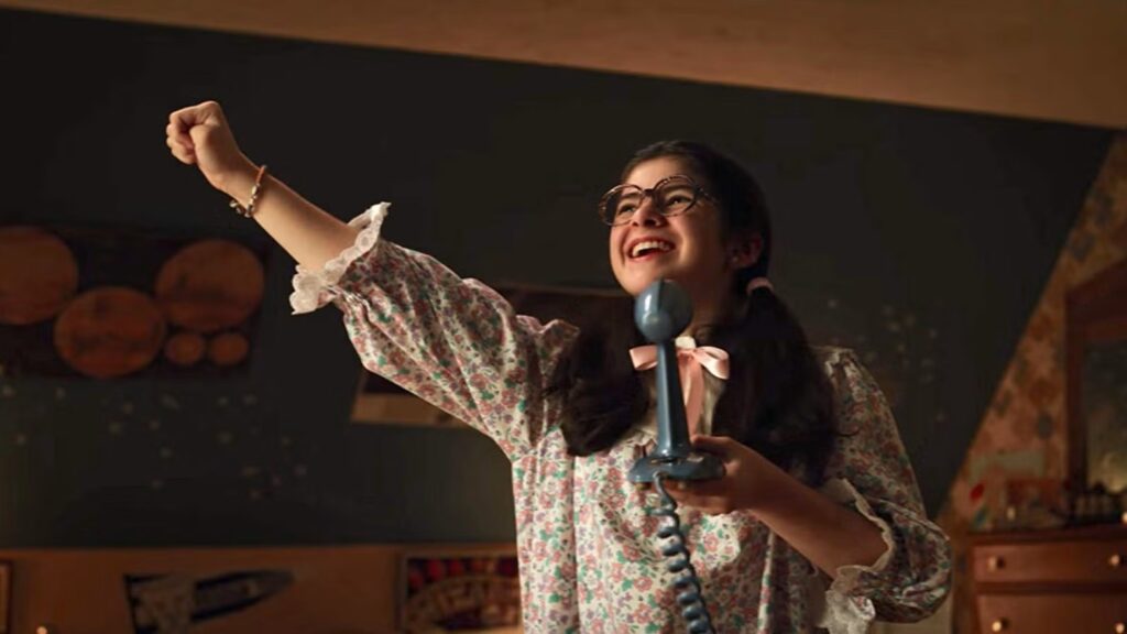 Suzie (Gabriella Pizzolo) enthusiastically singing into a radio microphone with her fist raised in the air, wearing glasses and appearing joyful.