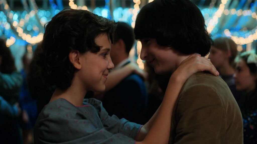 Eleven (Millie Bobby Brown) and Mike Wheeler (Finn Wolfhard) slow dancing at the Snow Ball, with romantic blue lighting and other couples in the background.
