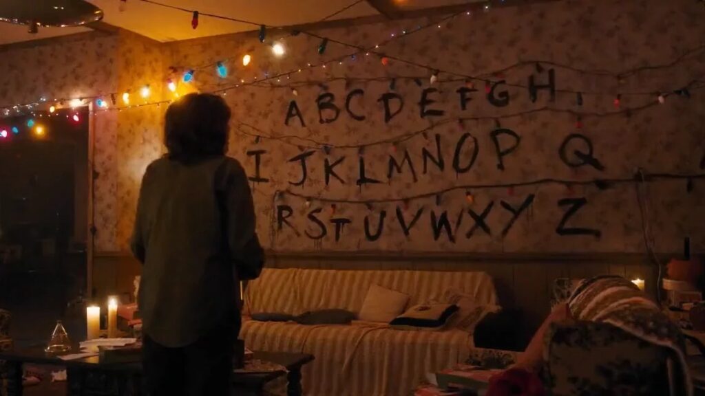 Joyce Byers (Winona Ryder) standing in her living room with Christmas lights strung on the wall behind her, with the alphabet painted on the wall for communication.