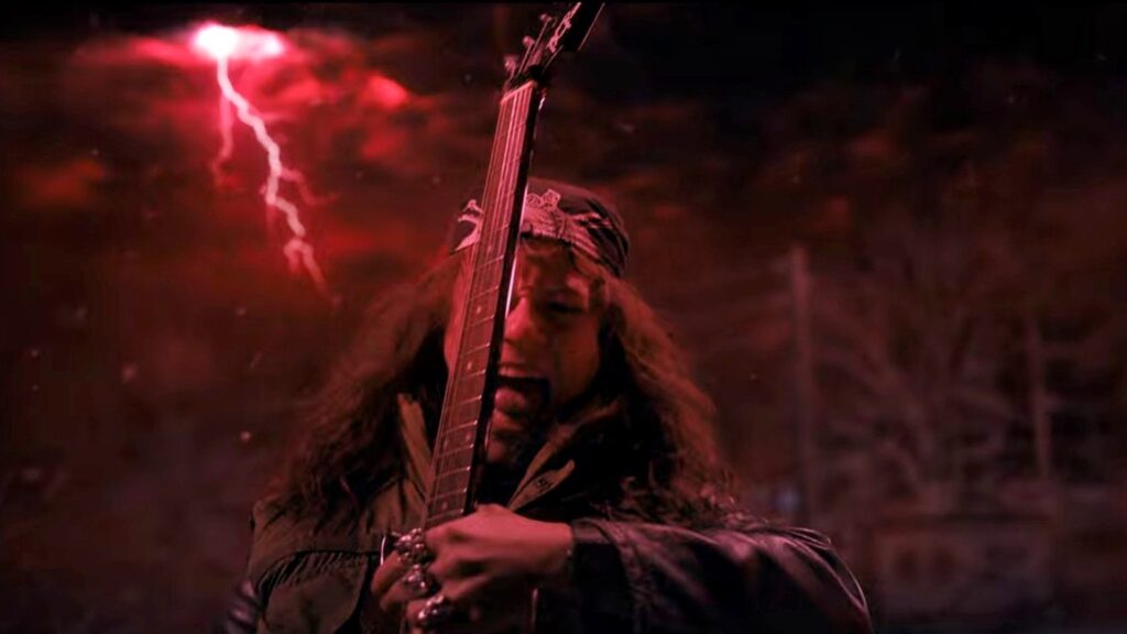 Eddie Munson (Joseph Quinn) playing electric guitar with long hair flowing, set against a dramatic red-tinted background with lightning, performing in the Upside Down.