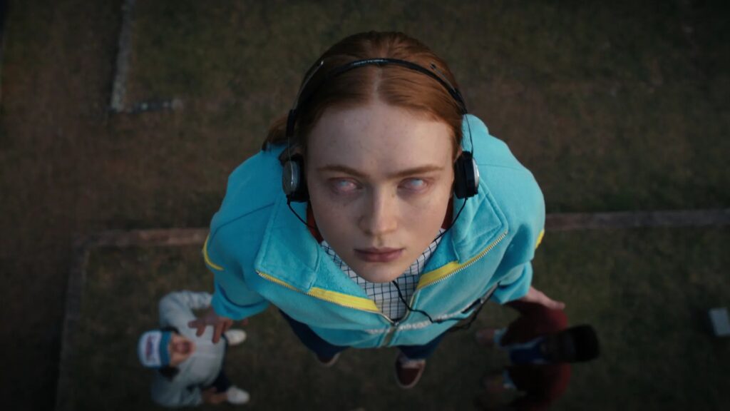Max Mayfield (Sadie Sink) wearing headphones and a blue winter jacket, looking up with an intense expression in a still from Stranger Things Season 4.