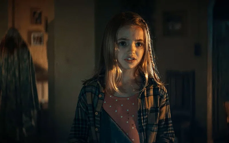 Nell Fisher as Kassie in Evil Dead Rise wearing plaid shirt standing in dimly lit apartment