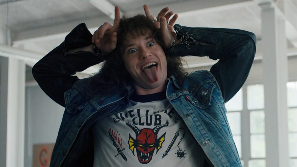 Eddie Munson making a playful face wearing his Hellfire Club t-shirt and denim jacket in Stranger Things
