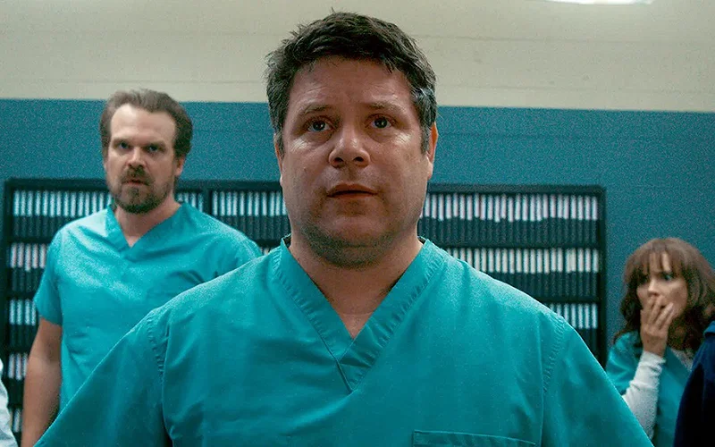Bob Newby (Sean Astin) in Hawkins Lab wearing scrubs in Stranger Things Season 2, Hopper (David Harbour) and Joyce (Winona Ryder) are visible in the background