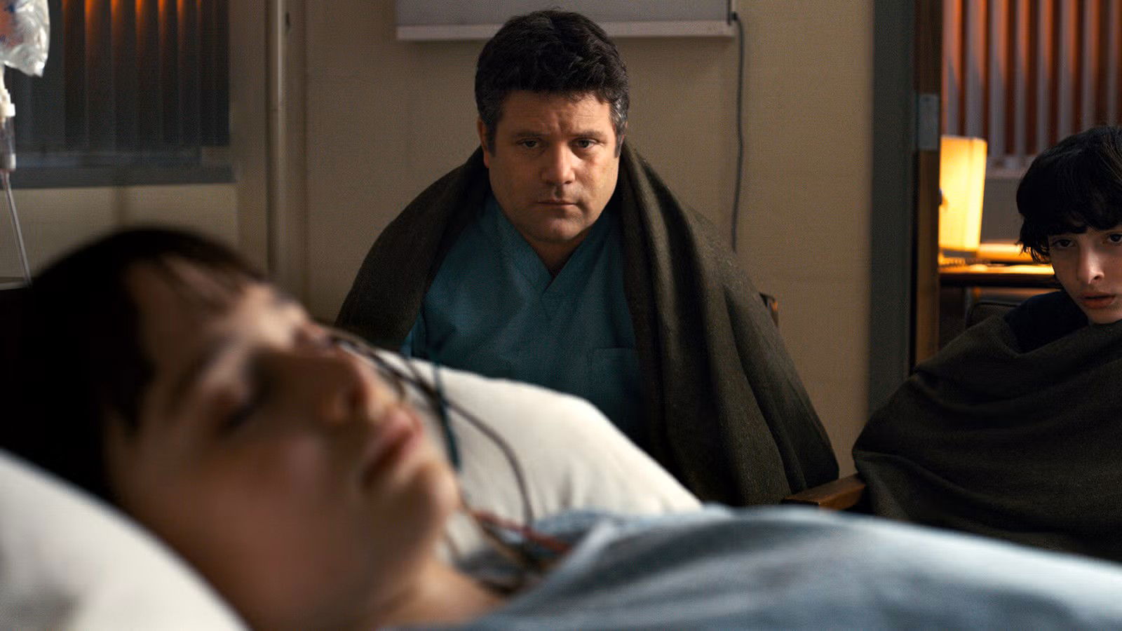 Bob Newby (Sean Astin) sitting on a hospital chair next to Mike Wheeler (Finn Wolfhard) on the side while Will Byers (Noah Schnapp) lies in bed in Stranger Things