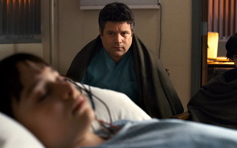 Bob Newby (Sean Astin) sitting on a hospital chair next to Mike Wheeler (Finn Wolfhard) on the side while Will Byers (Noah Schnapp) lies in bed in Stranger Things