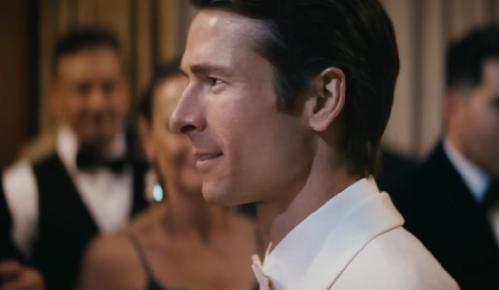Glen Powell wearing a white suit in How to Make a Killing