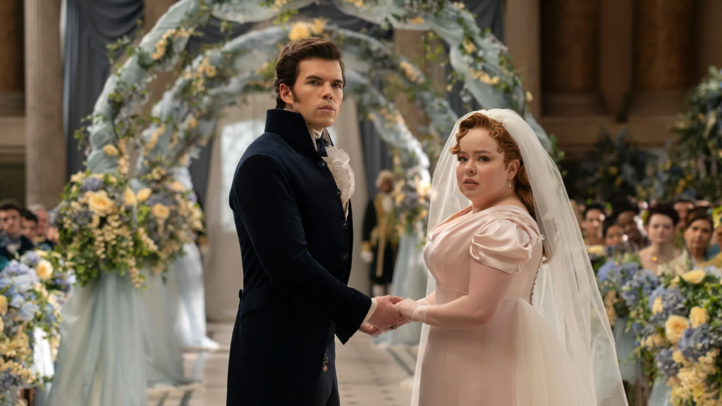 Luke Newton as Colin Bridgerton wearing a dark long tux standing on left with a wedding aisle in the background and standing opposite Nicola Coughlan as Penelope Featherington who is wearing a wedding gown and veil in Bridgerton | Credit: Netflix