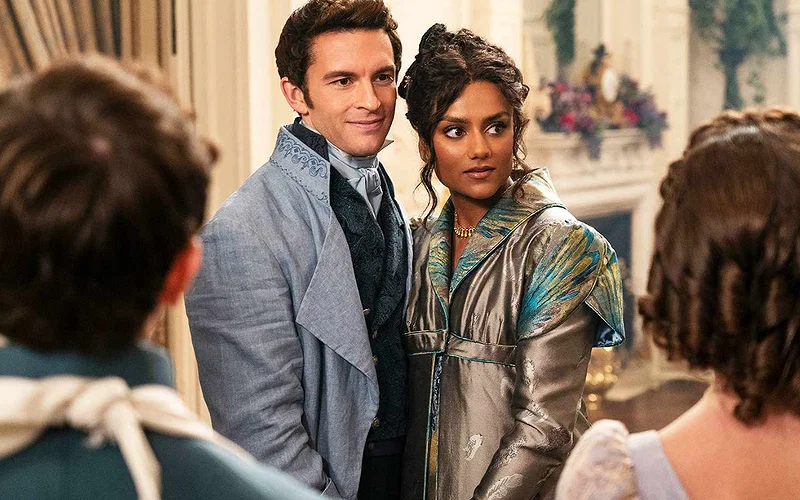 Jonathan Bailey as Anthony Bridgerton wearing a washed blue suit and standing next to Simone Ashley who plays Kate and wears a silk printed grey and blue gown and looks away from the camera in Bridgerton | Credit: Netflix