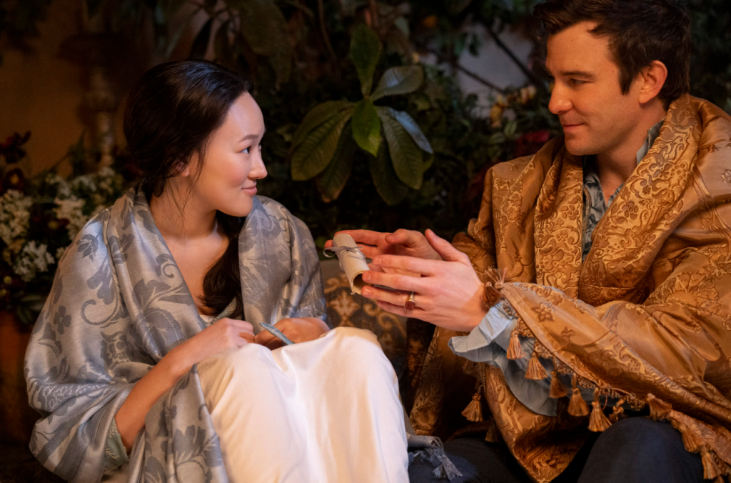 Benedict Bridgerton (Luke Thompson) on the right has a beige shawl draped around him and offers a scroll to Sophie Baek (Yerin Ha) who is on his left and has a great shawl around her and is smiling. Credit: Netflix