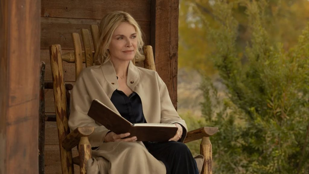 Michelle Pfeiffer sitting in a wooden rocking chair on a cabin porch reading a book