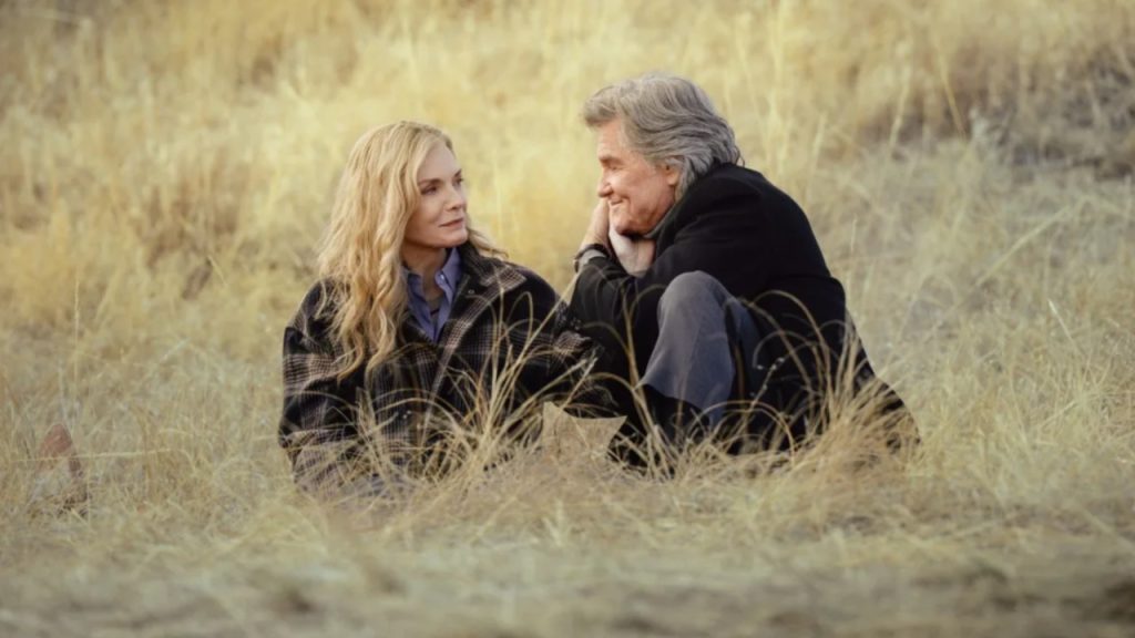 Alt Text: Michelle Pfeiffer and Kurt Russell sitting together in a dry grass field in a scene from The Madison