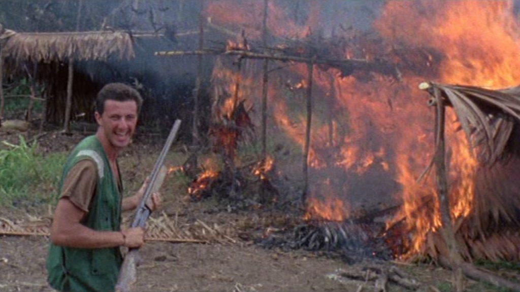 A man laughing and in background a house caught fire in Cannibal Holocaust