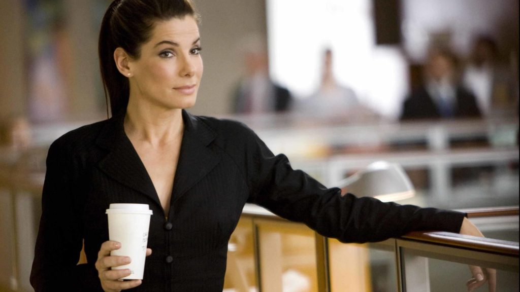 Sandra Bullock holding a cup of takeaway coffee stands in a modern office setting

