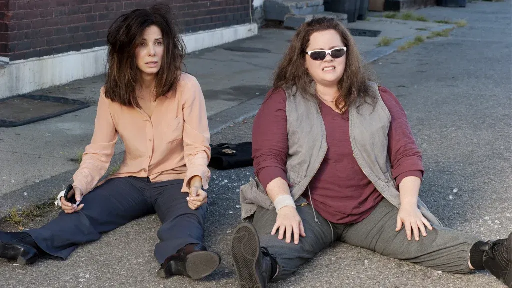 Sandra Bullock and Melissa McCarthy sit on a street, looking shaken and uncomfortable. 