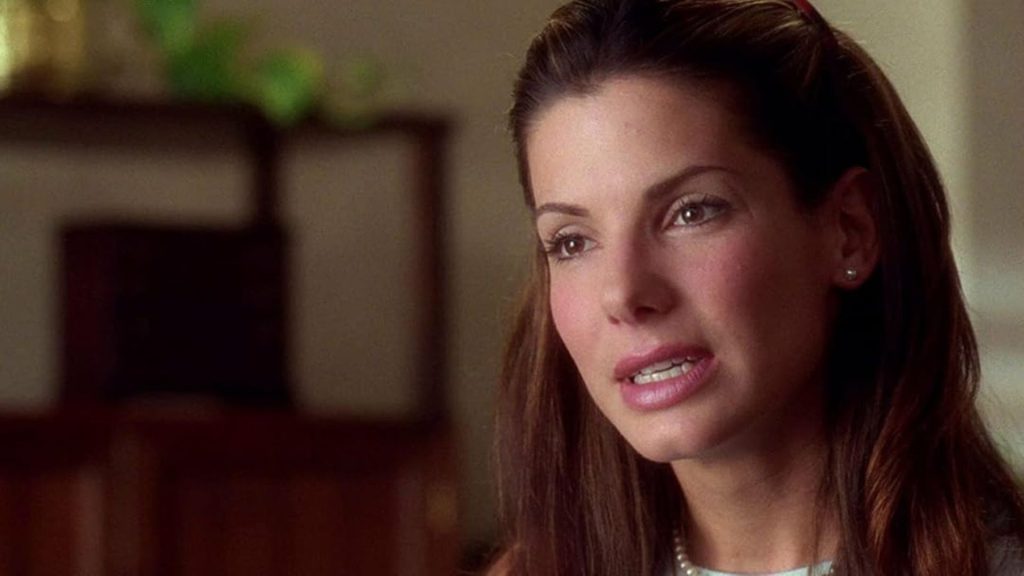 Sandra Bullock with brown hair saying something in softly-lit indoors. The background is blurred and warm-toned