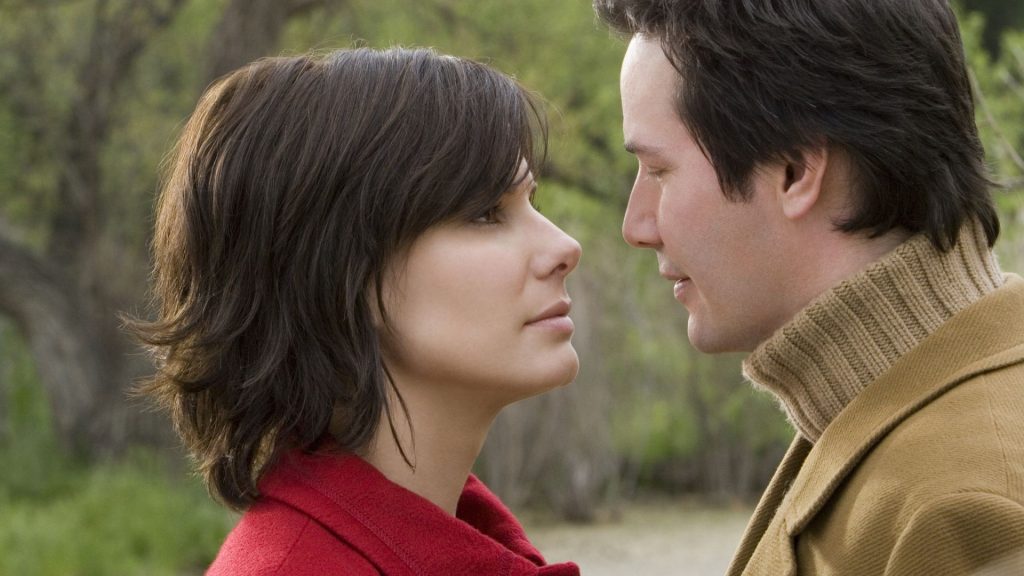 Sandra Bullock and Keanu Reeves stand very close outdoors, gazing at each other with an intimate expression.