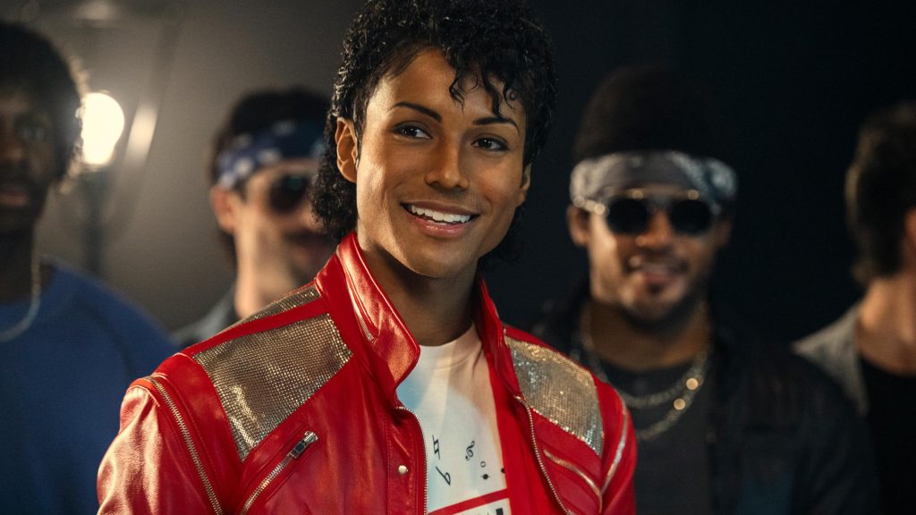 Actor portraying Michael Jackson in the biopic Michael, wearing a red leather jacket backstage, performing before a packed stadium crowd, and preparing to put on sunglasses under stage lights.