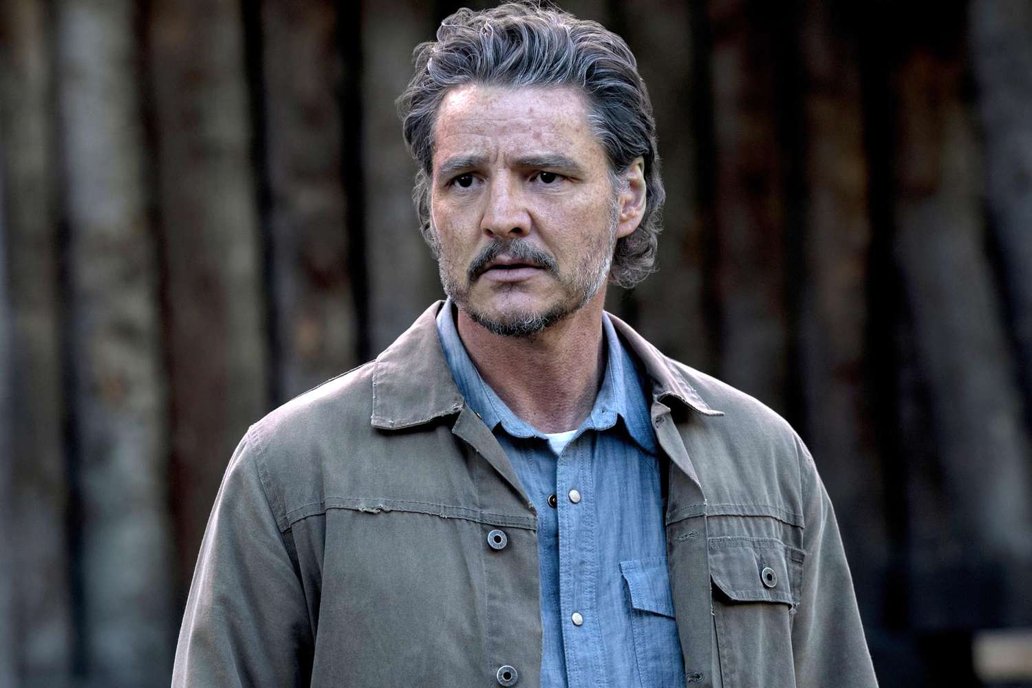 Pedro Pascal as a man with graying hair and facial stubble stands outdoors in front of blurred wooden structures, wearing a blue shirt and light brown jacket. His concerned expression suggests a tense or emotional moment.