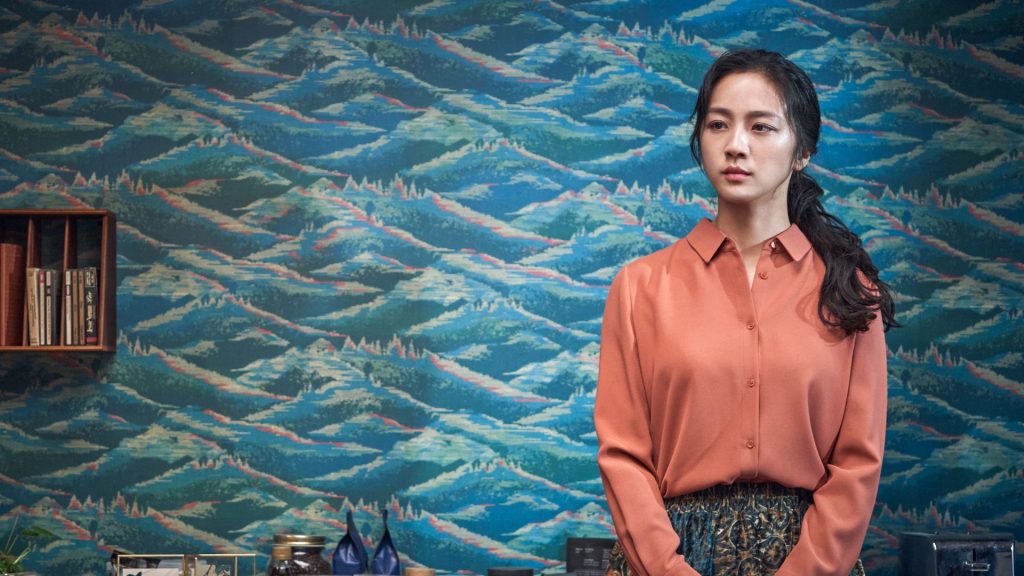A woman stands indoors against a vivid blue patterned wall, wearing a coral blouse and patterned skirt. She looks thoughtfully to the side, while shelves, books, and decorative objects add a stylish and composed atmosphere to the room
