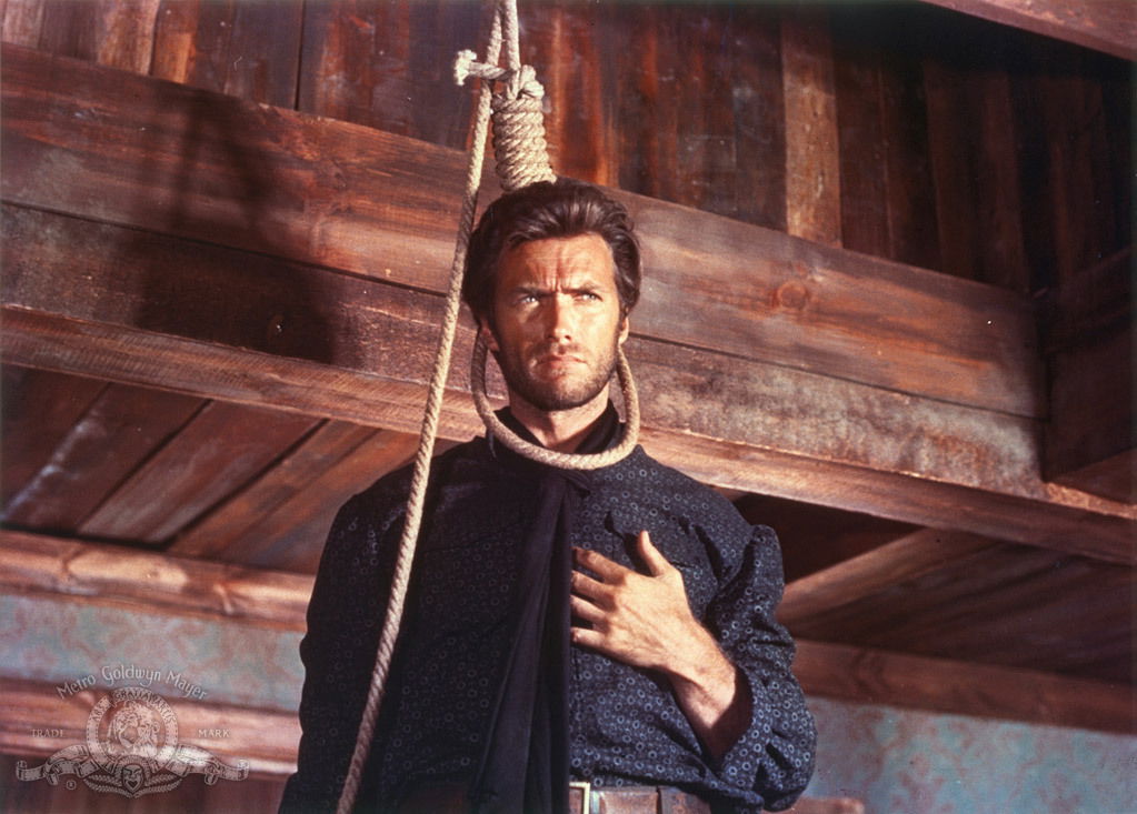 A stern-looking man stands beneath a wooden beam with a rope noose hanging around his neck, one hand pressed to his chest. Warm light falls across the rustic interior, creating a tense Old West standoff atmosphere.