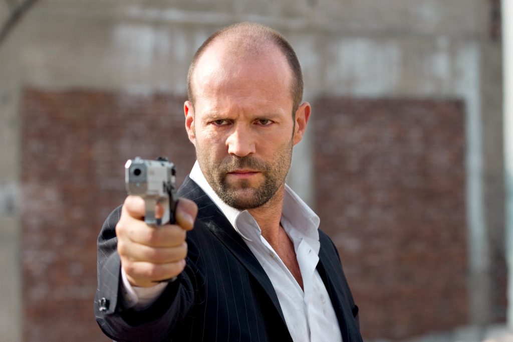 Jason Statham in a dark suit points a handgun directly toward the camera, staring intensely, with a blurred brick wall in the background.
