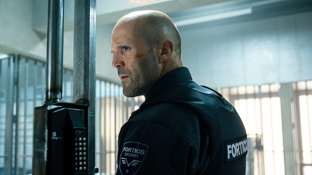Jason Statham in a dark security uniform turns sharply inside a secure facility, with metal bars and bright overhead lighting in the background.