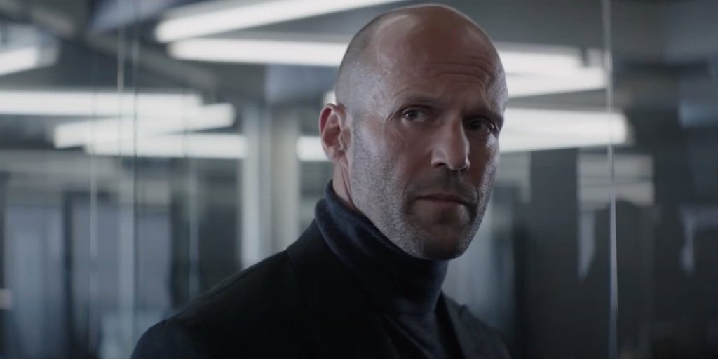 Jason Statham wearing a dark turtleneck and coat looks sideways inside a modern glass-walled room with bright overhead lights.