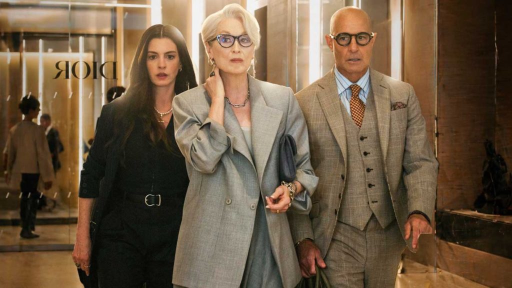 Three sharply dressed professionals walk through a luxury fashion building entrance, with a poised woman in a grey suit at the center flanked by a younger woman and a man in a tailored suit.