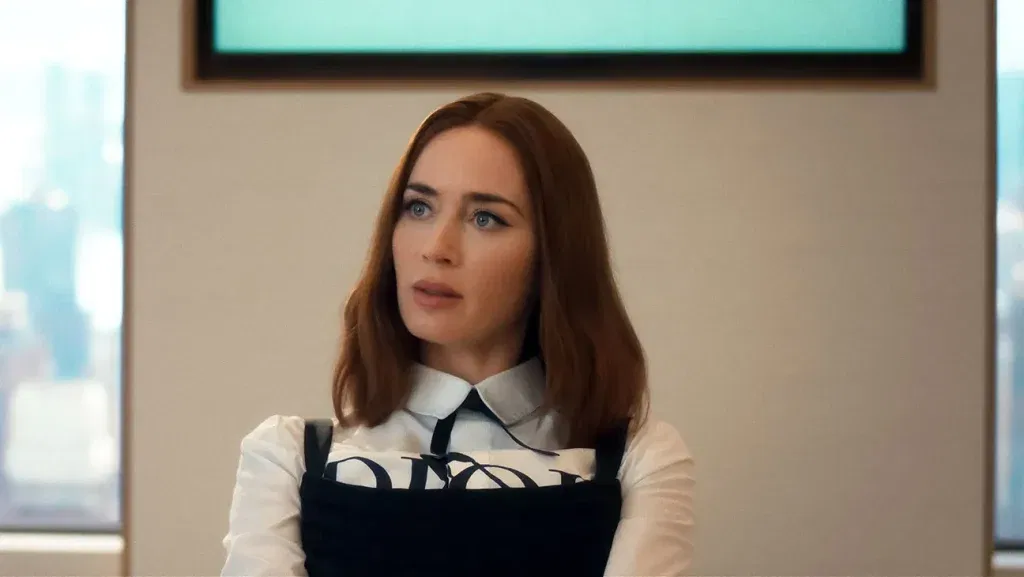 A woman with shoulder-length auburn hair sits indoors wearing a structured black dress over a white collared shirt, looking slightly surprised or attentive during a conversation.