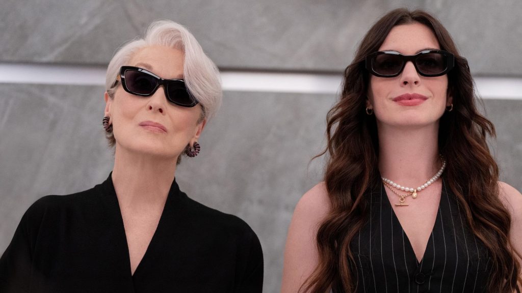 Two stylish women wearing black outfits and sunglasses stand side by side, looking composed and confident against a neutral backdrop.