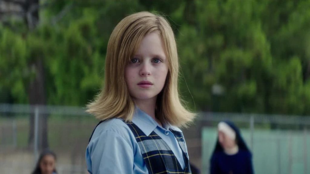 A young girl stares ahead with an unsettling expression while standing outdoors in a school uniform.