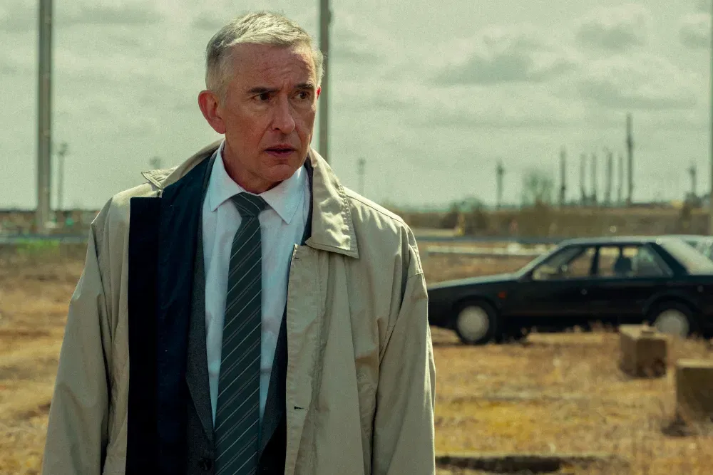 A grey-haired man wearing a beige trench coat and striped tie stands outdoors in a dry industrial area, looking anxiously to one side while an old dark-colored car sits behind him under a cloudy sky.