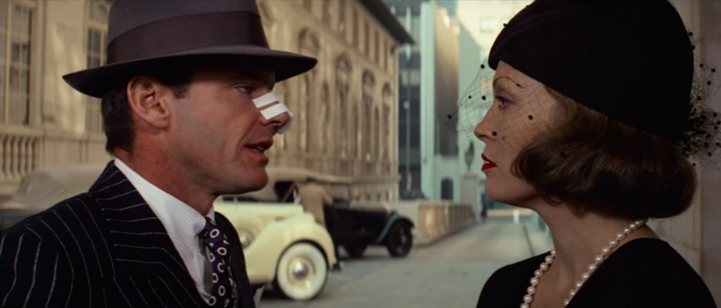 A man in a pinstripe suit and fedora with a bandage across his nose stands face-to-face with a woman wearing a black hat and pearl necklace on a city street in a scene from Chinatown.