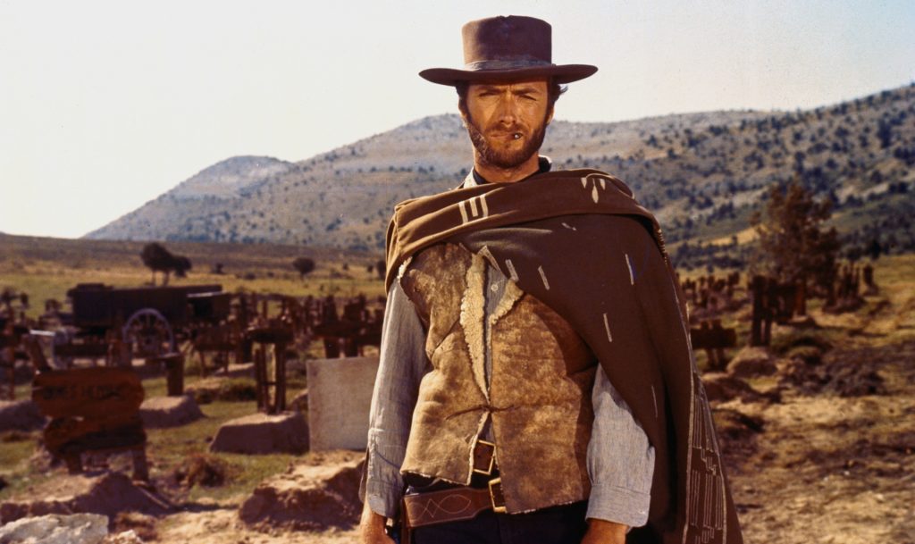Clint Eastwood stands in a dusty cemetery wearing a cowboy hat, poncho, and holster while staring ahead in a scene from The Good, the Bad and the Ugly.