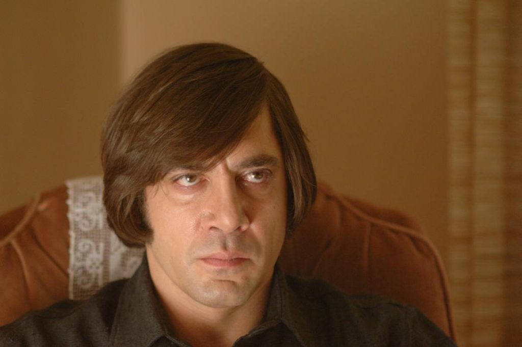 A man with a bowl haircut and an intense blank expression sits staring forward in a dimly lit room in a scene from No Country for Old Men.