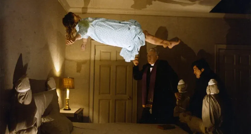 A young girl levitates above a bed inside a dimly lit bedroom while two priests perform an exorcism in a terrifying scene from The Exorcist.
