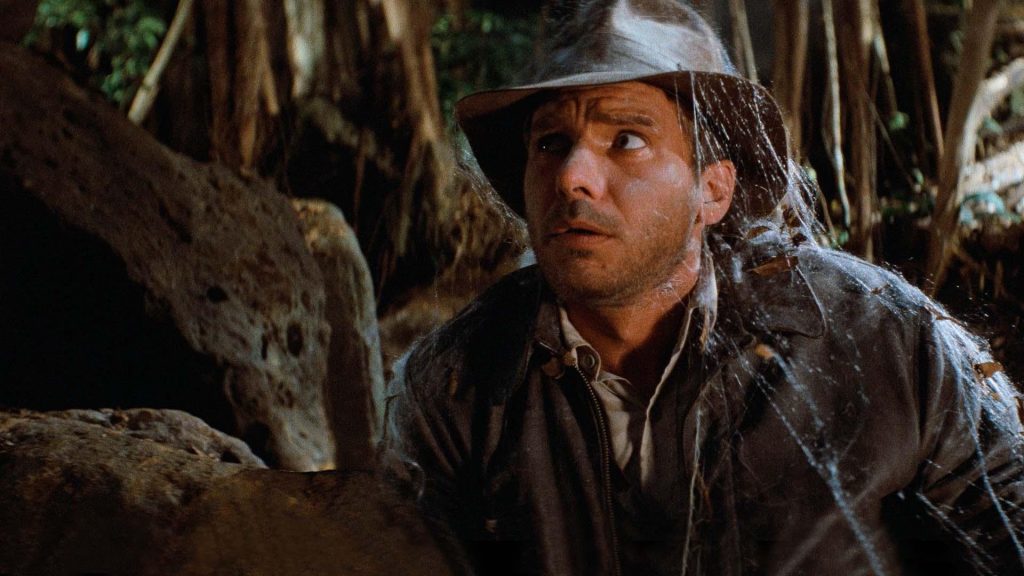 Harrison Ford as Indiana Jones crouches nervously inside a dark jungle setting while wearing his iconic fedora and leather jacket in Raiders of the Lost Ark.