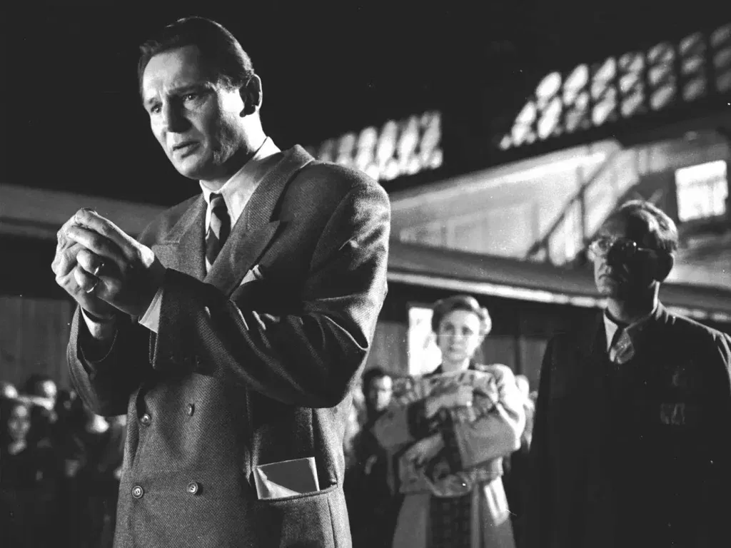 Liam Neeson as Oskar Schindler stands in a crowded black-and-white scene while examining an object in his hands in Schindler’s List.