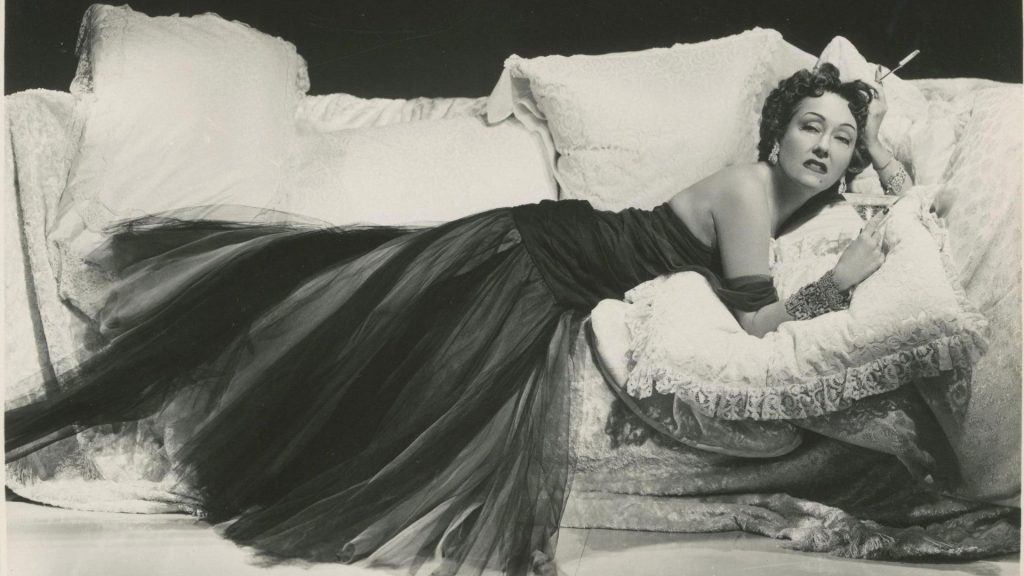 Gloria Swanson as Norma Desmond reclines dramatically on an ornate couch in a flowing dark gown while holding a cigarette in a black-and-white publicity image from Sunset Boulevard.