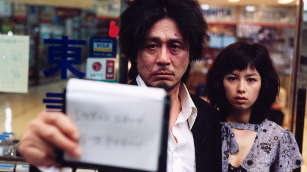 A disheveled man with bruises and messy hair stares blankly ahead while holding up a small object, with a young woman standing behind him in a scene from Oldboy.