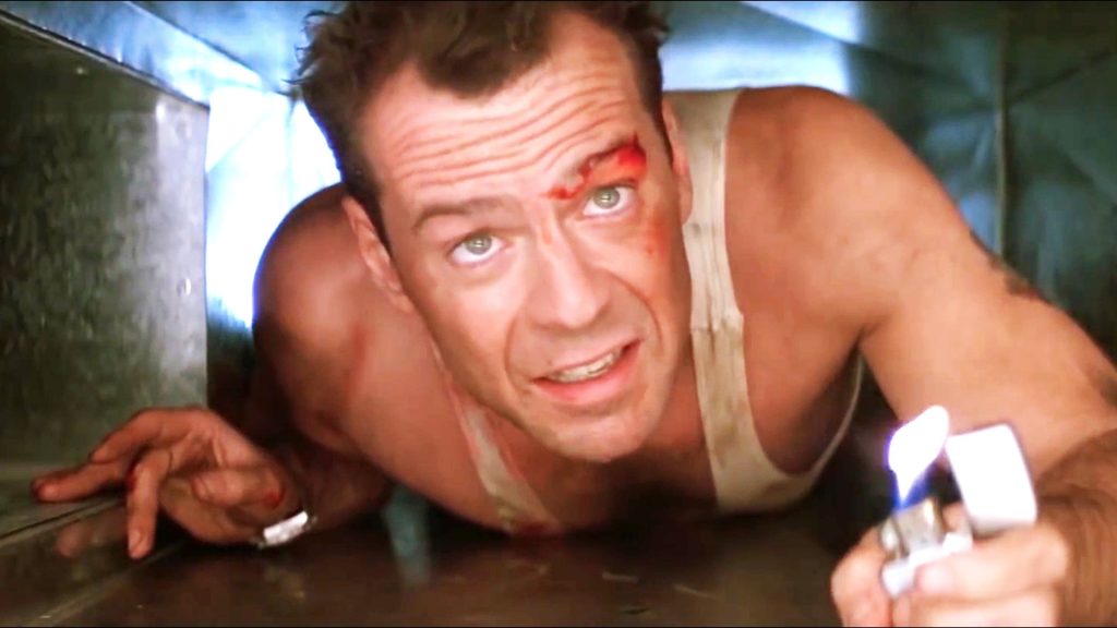 Bruce Willis as John McClane crawls through a narrow air vent while injured and holding a lighter in a tense scene from Die Hard.