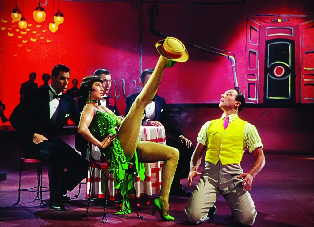 A woman in a sparkling green dress performs a high kick in a brightly colored nightclub while a man in a yellow vest kneels beside her in a musical number from Singin’ in the Rain.
