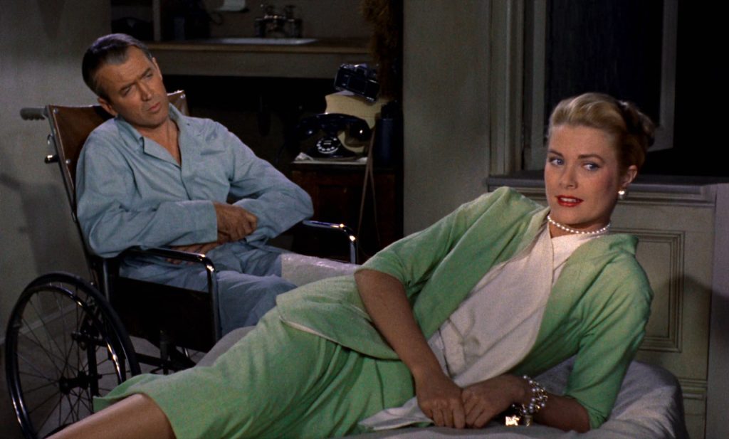 Grace Kelly reclines elegantly on a couch while James Stewart sits watching from a wheelchair in a scene from Alfred Hitchcock’s Rear Window.