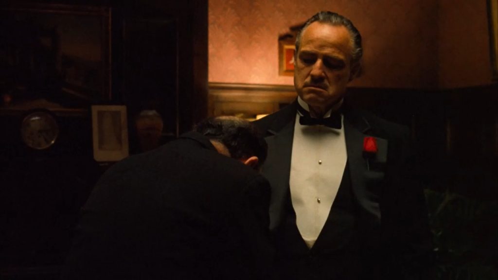 Marlon Brando as Don Vito Corleone stands solemnly in a dark room wearing a tuxedo while another man bows before him in a scene from The Godfather.