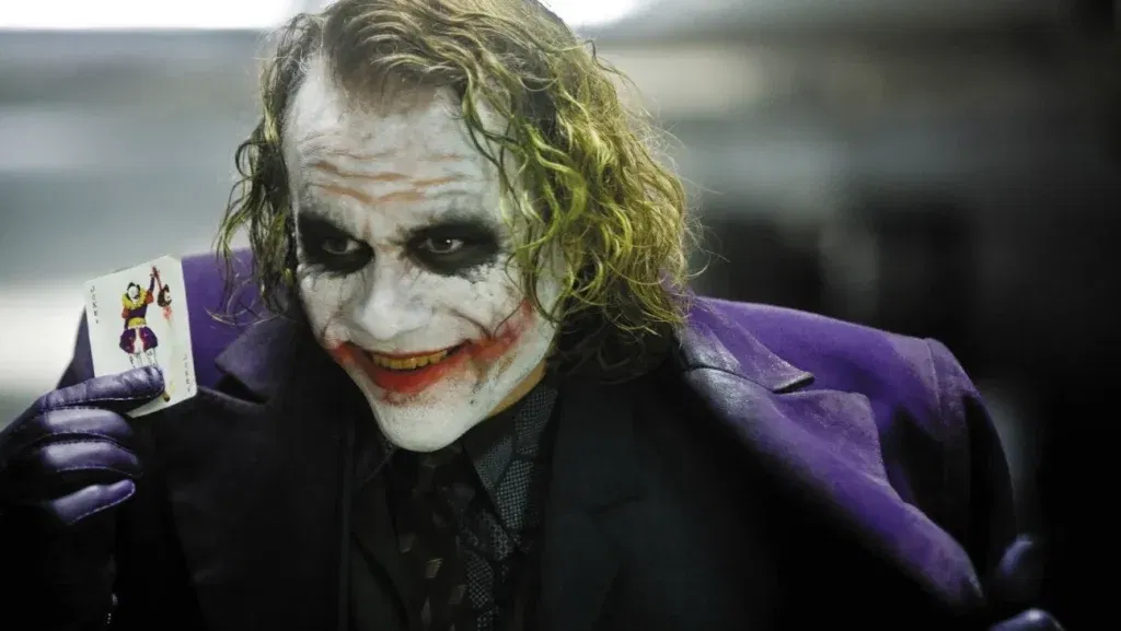 Heath Ledger as the Joker, wearing smeared clown makeup and a purple coat, grins while holding up a Joker playing card in The Dark Knight.